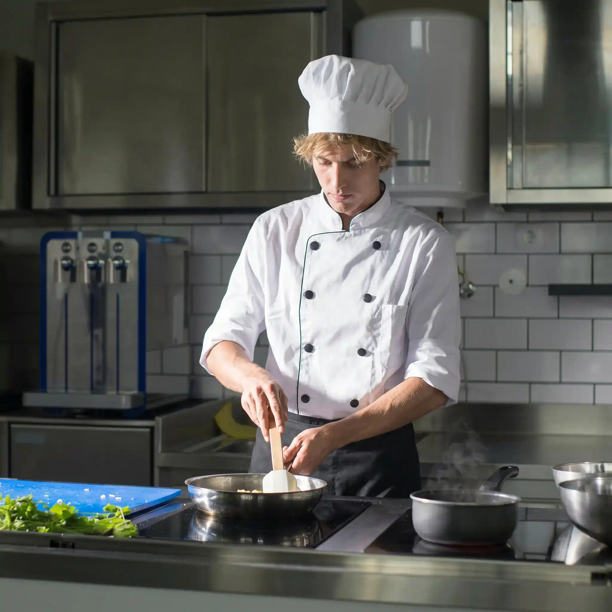 Chef in a professional kitchen stirring a pot on a stove.